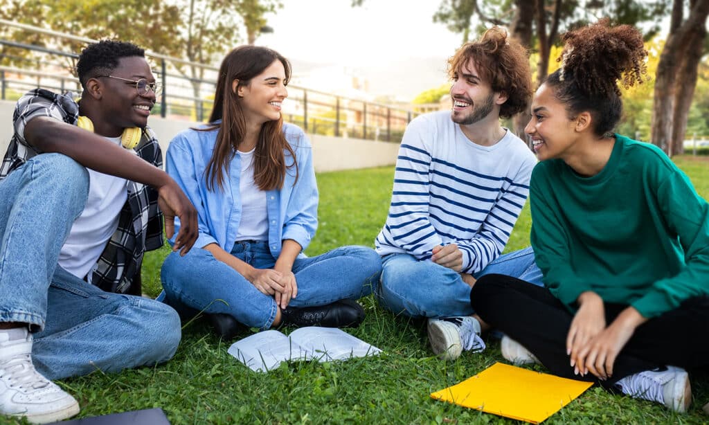 A group of four friends sit in the grass smiling and reading their Bible at a park.