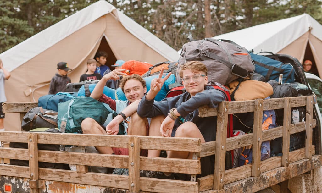 Two boys sit in the back of a wooden bed truck full of camping gear, smiling at the camera