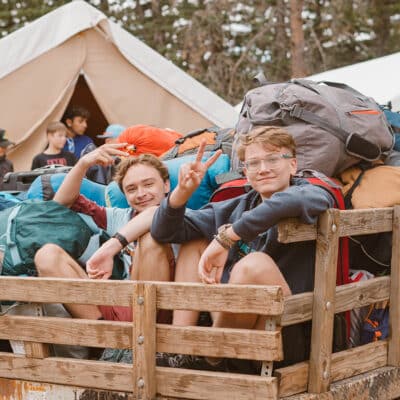 Two boys sit in the back of a wooden bed truck full of camping gear, smiling at the camera