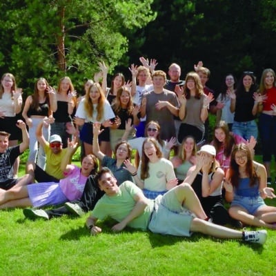 A large group of young adults poses together on a sunny lawn, smiling and waving enthusiastically at the camera.