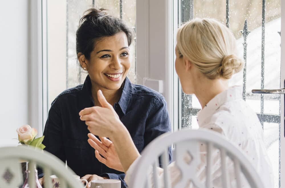 Two women sit at a small table by a window, engaged in a warm and friendly conversation.