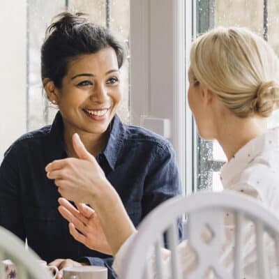 Two women sit at a small table by a window, engaged in a warm and friendly conversation.