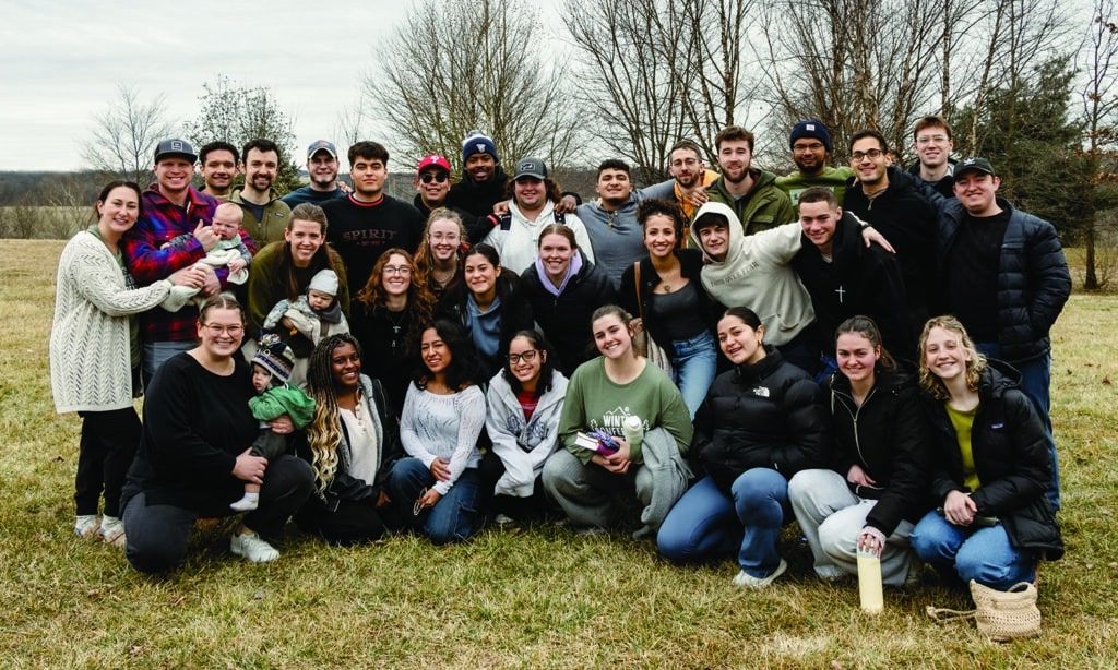 052025 - Article - Web A large group of young adults and a few small children pose together outdoors on a cloudy day, smiling warmly at the camera. They are bundled up in winter and fall clothing, standing on a grassy field with bare trees in the background.