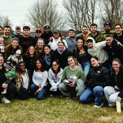 A large group of young adults and a few small children pose together outdoors on a cloudy day, smiling warmly at the camera. They are bundled up in winter and fall clothing, standing on a grassy field with bare trees in the background.