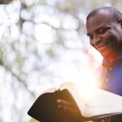 A man wearing a blue polo shirt smiles as he reads an open Bible outdoors. Sunlight shines brightly from the pages, and a soft-focus background of trees creates a peaceful atmosphere.