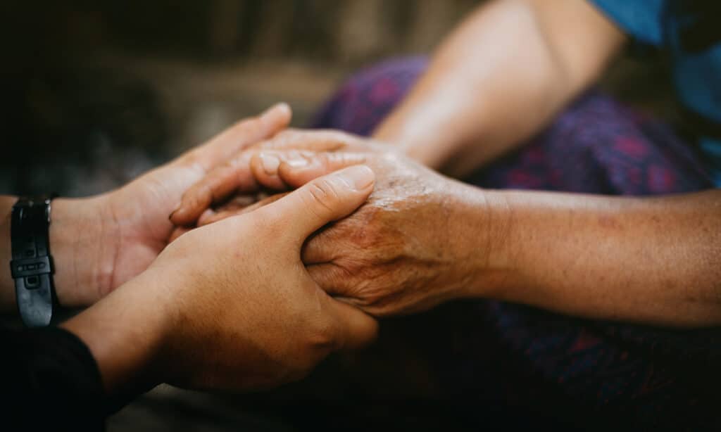 Two people gently hold hands in a comforting gesture. One pair of hands appears older and weathered, while the other pair is younger.