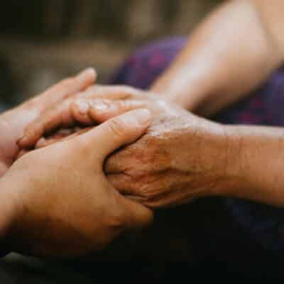 Two people gently hold hands in a comforting gesture. One pair of hands appears older and weathered, while the other pair is younger.