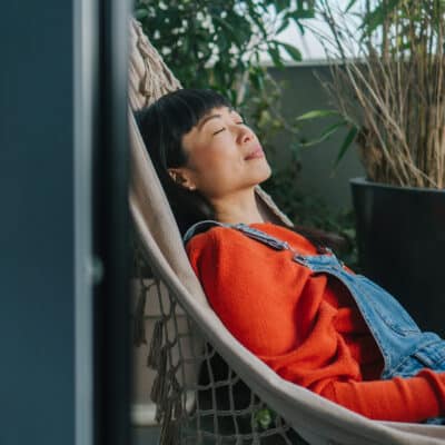 A woman relaxes in a hammock on a balcony surrounded by potted plants. She is wearing a red sweater and denim overalls, with her eyes closed and a peaceful expression. She holds a closed book in one hand, and soft natural light fills the scene.