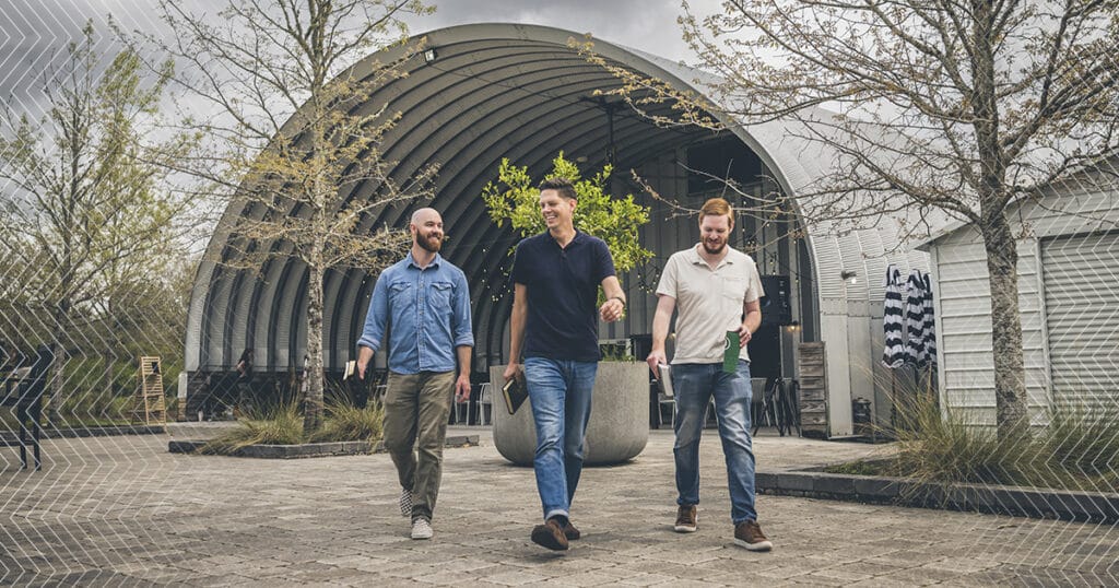 Three men walk outdoors in front of a modern, arched metal building, each holding a Bible and smiling in conversation, surrounded by leafless trees and cloudy skies.