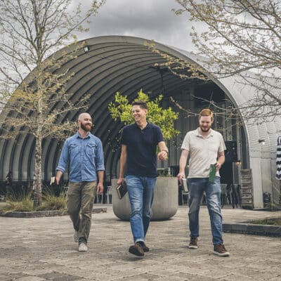 Three men walk outdoors in front of a modern, arched metal building, each holding a Bible and smiling in conversation, surrounded by leafless trees and cloudy skies.