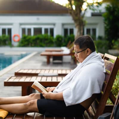 A man relaxes on a poolside lounge chair, wrapped in a white towel, reading a book with focused attention at a serene resort setting.