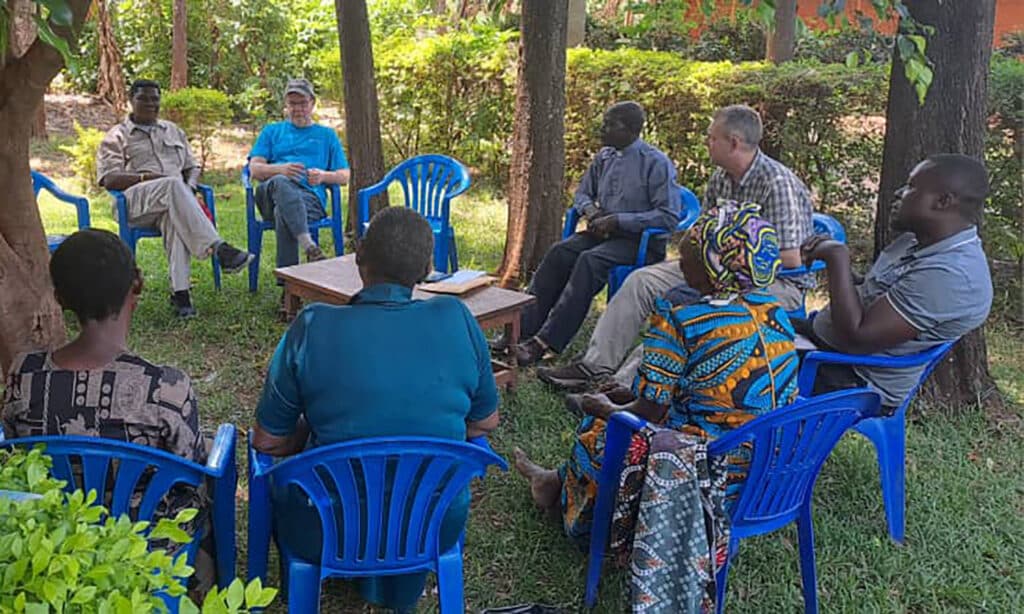 A diverse group of people gather in a circle of blue chairs under trees for a discussion, with a Bible open on a small wooden table in the center.
