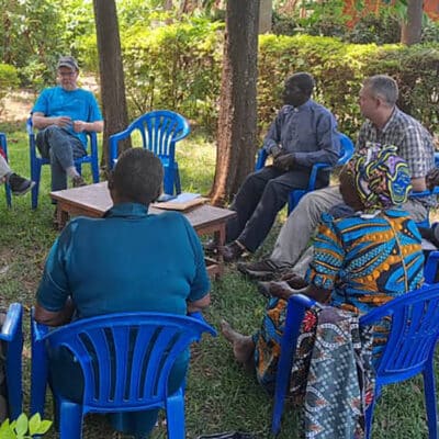 A diverse group of people gather in a circle of blue chairs under trees for a discussion, with a Bible open on a small wooden table in the center.