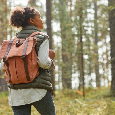 Back view portrait of young African-American woman with backpack enjoying praying and fasting in forest lit by sunlight.