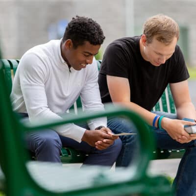 Two young men sit on a bench with Bibles in hand praying together.