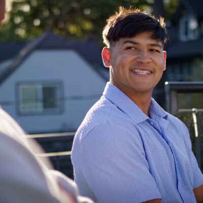 A young man smiling warmly during a conversation on an outdoor patio, with houses and trees in the background on a sunny day.