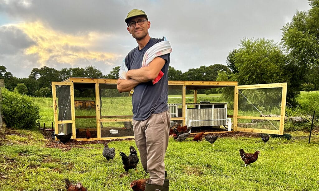 A man in a T-shirt and long pants stands in a field with a chicken coup behind him and chickens walking around him.