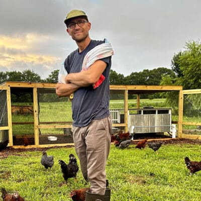 A man in a T-shirt and long pants stands in a field with a chicken coup behind him and chickens walking around him.