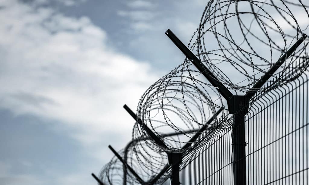 The top of a jailyard fence with barped wire. The image is taken from the bottom of the fence with the blue sky in the background.