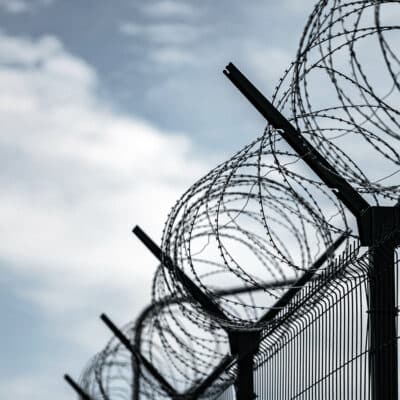 The top of a jailyard fence with barped wire. The image is taken from the bottom of the fence with the blue sky in the background.