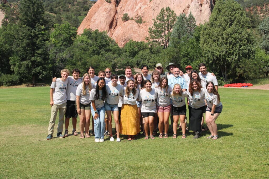 A group of people stand, posed for a picture on a grass field with a big rock structure behind them in the distance.