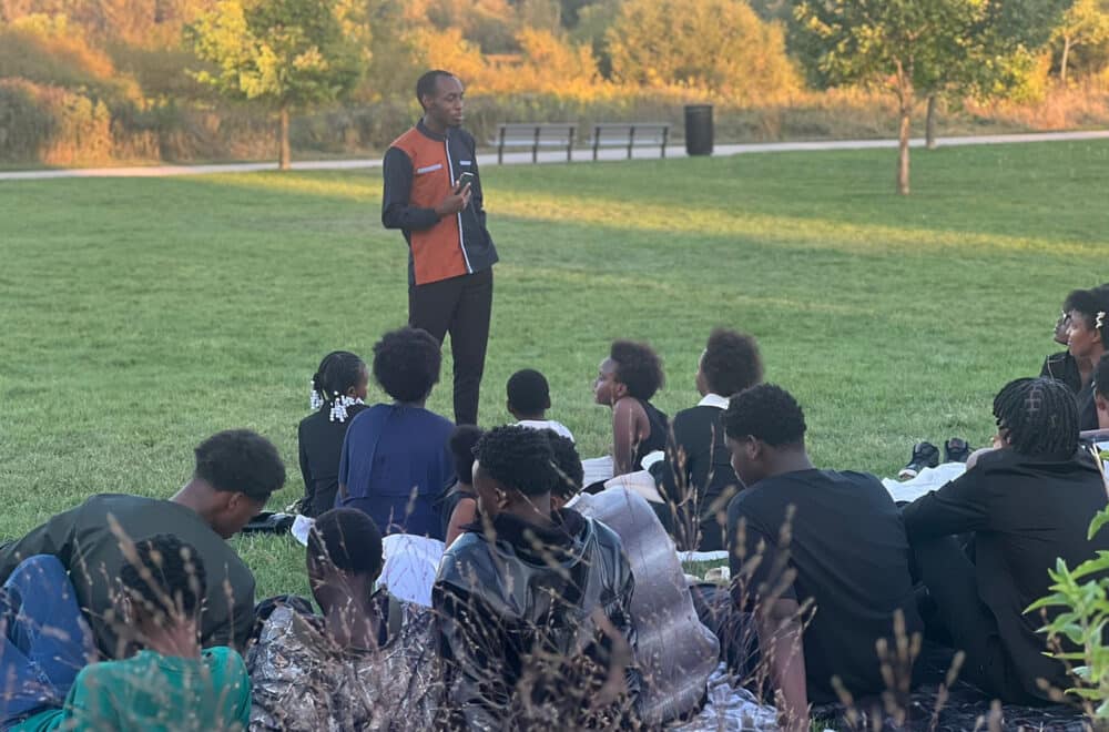 A man stands outdoors on a grassy field speaking to a seated group of children and teens. The group listens attentively as the sun sets behind them, casting warm light over the trees in the background.
