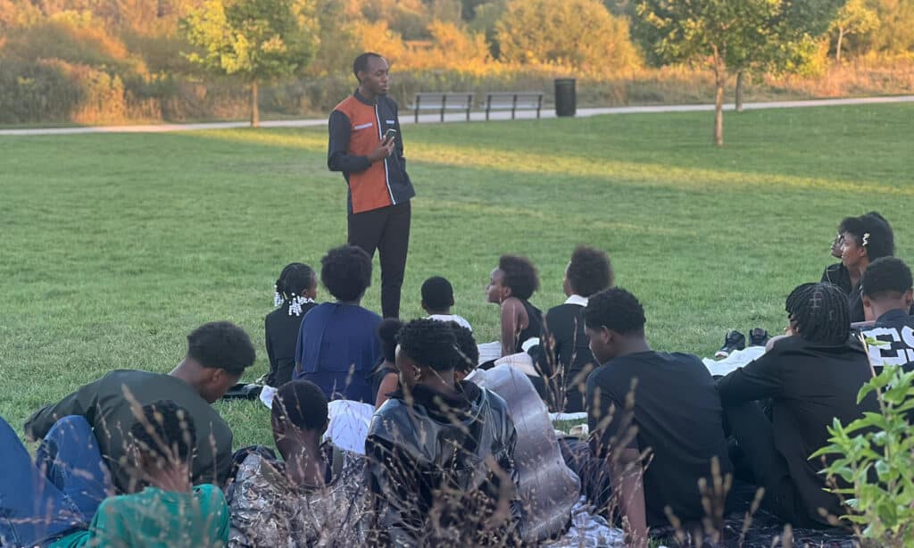 A man stands outdoors on a grassy field speaking to a seated group of children and teens. The group listens attentively as the sun sets behind them, casting warm light over the trees in the background.