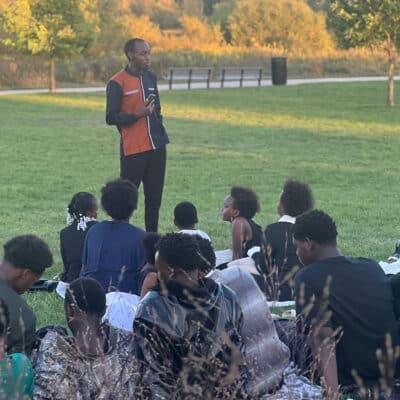 A man stands outdoors on a grassy field speaking to a seated group of children and teens. The group listens attentively as the sun sets behind them, casting warm light over the trees in the background.