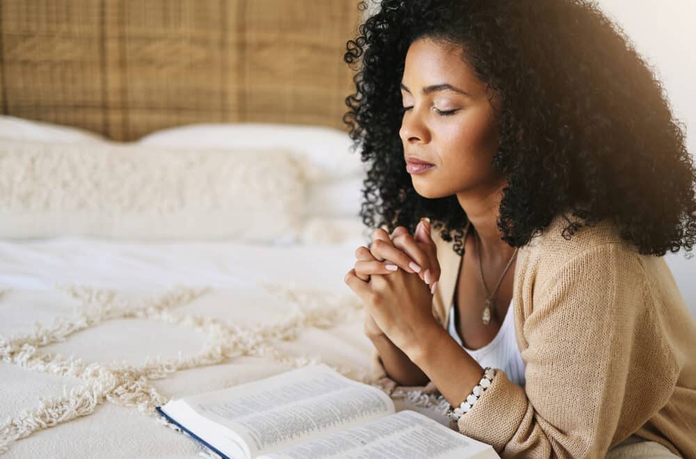 A woman sits on a bed with her hands clasped in prayer, eyes closed, beside an open Bible resting on the blanket.