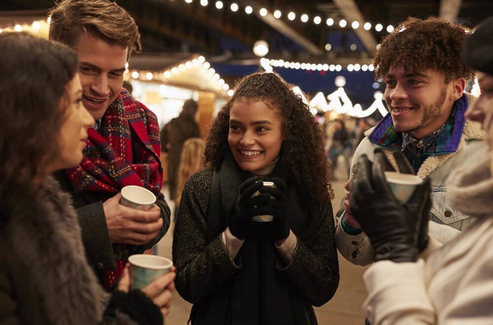 A group of friends smiles and talks while holding hot drinks at an outdoor market lit by string lights at night.