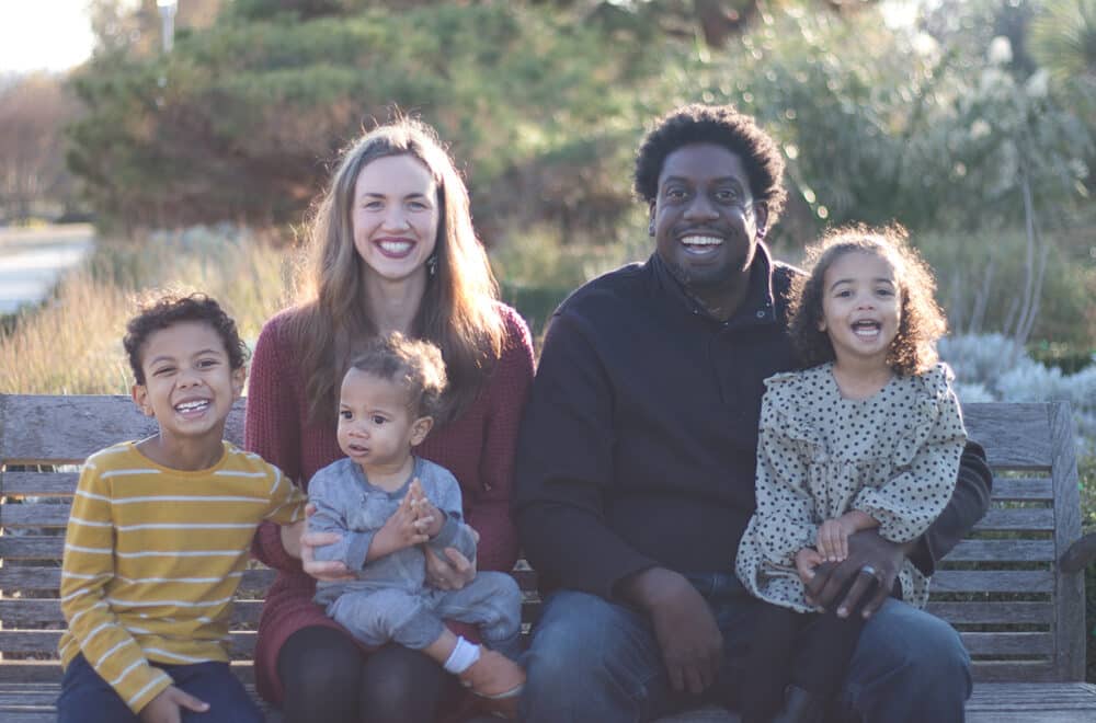 A family of five sits together on an outdoor bench, smiling and laughing in the sunlight.