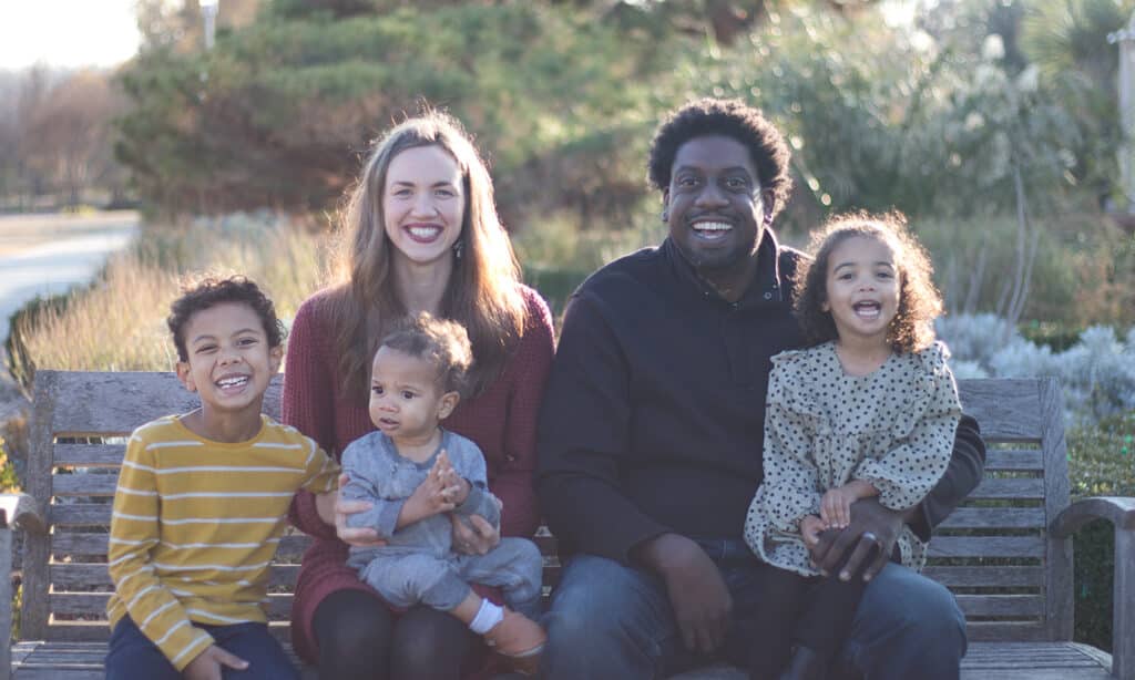 A family of five sits together on an outdoor bench, smiling and laughing in the sunlight.
