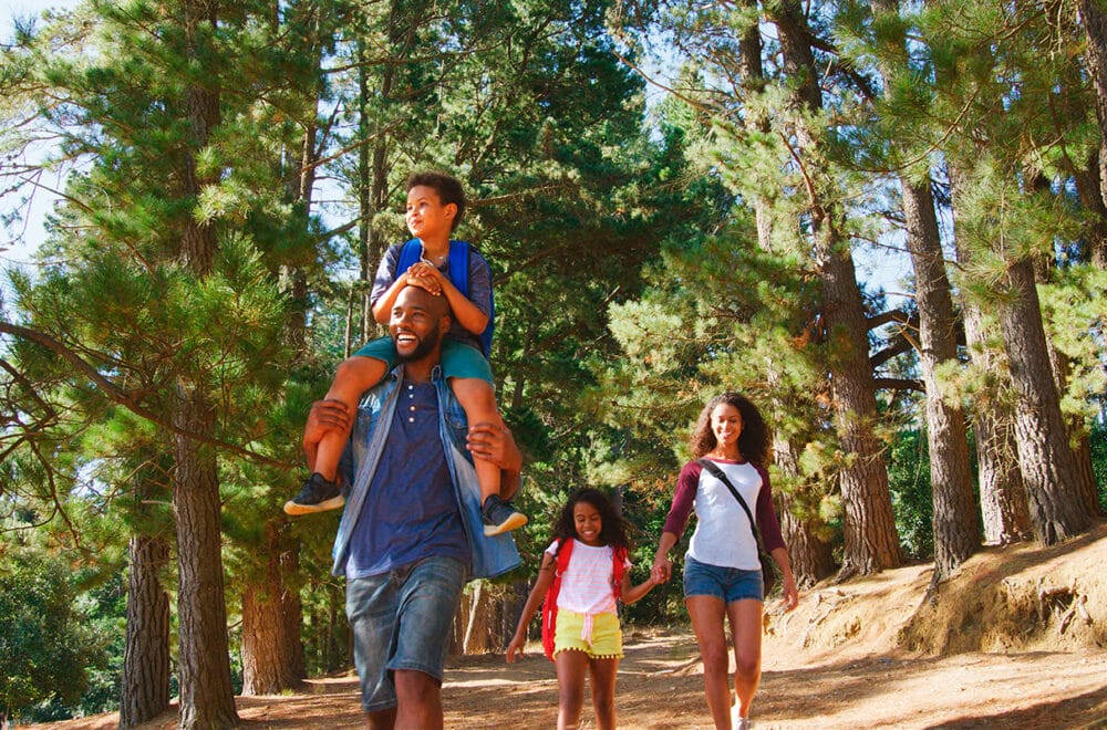 A family walks through a forest trail together, with the father carrying a child on his shoulders and the mother and daughter walking beside them.