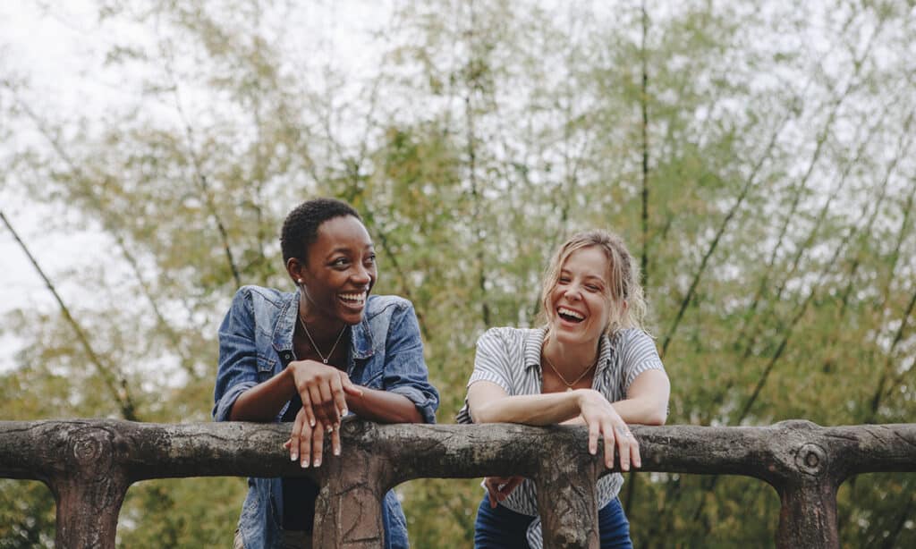 Two women leaning against a wooden handrail in the outdoors. They are smiling and laughing with each other.