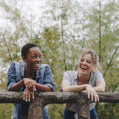 Two women leaning against a wooden handrail in the outdoors. They are smiling and laughing with each other.