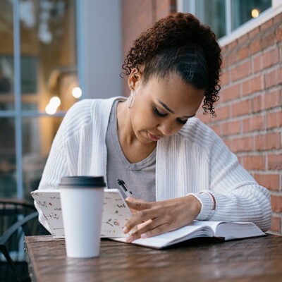 A mixed race women reads a large Bible / textbook while seated at table outdoors at a coffee shop.