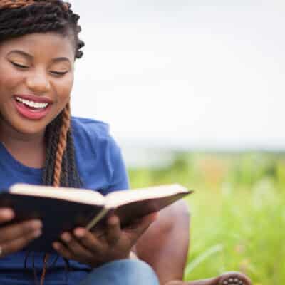 An African-American woman smiling while reading the Bible outside in a field.